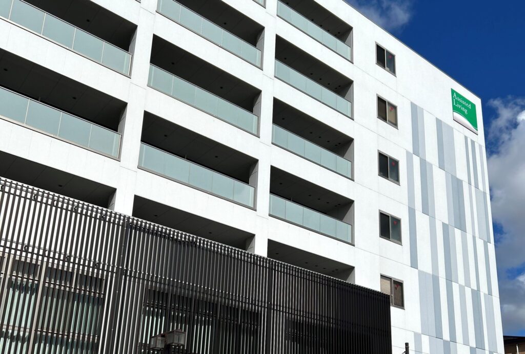 白を基調とした現代的な介護施設の建物外観。複数階のバルコニーと緑のロゴが見えます。