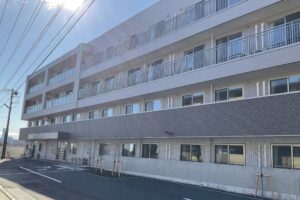 介護施設の4階建て建物外観。昼間の全体像が明確に映り、複数の居室窓と入口が見える。
